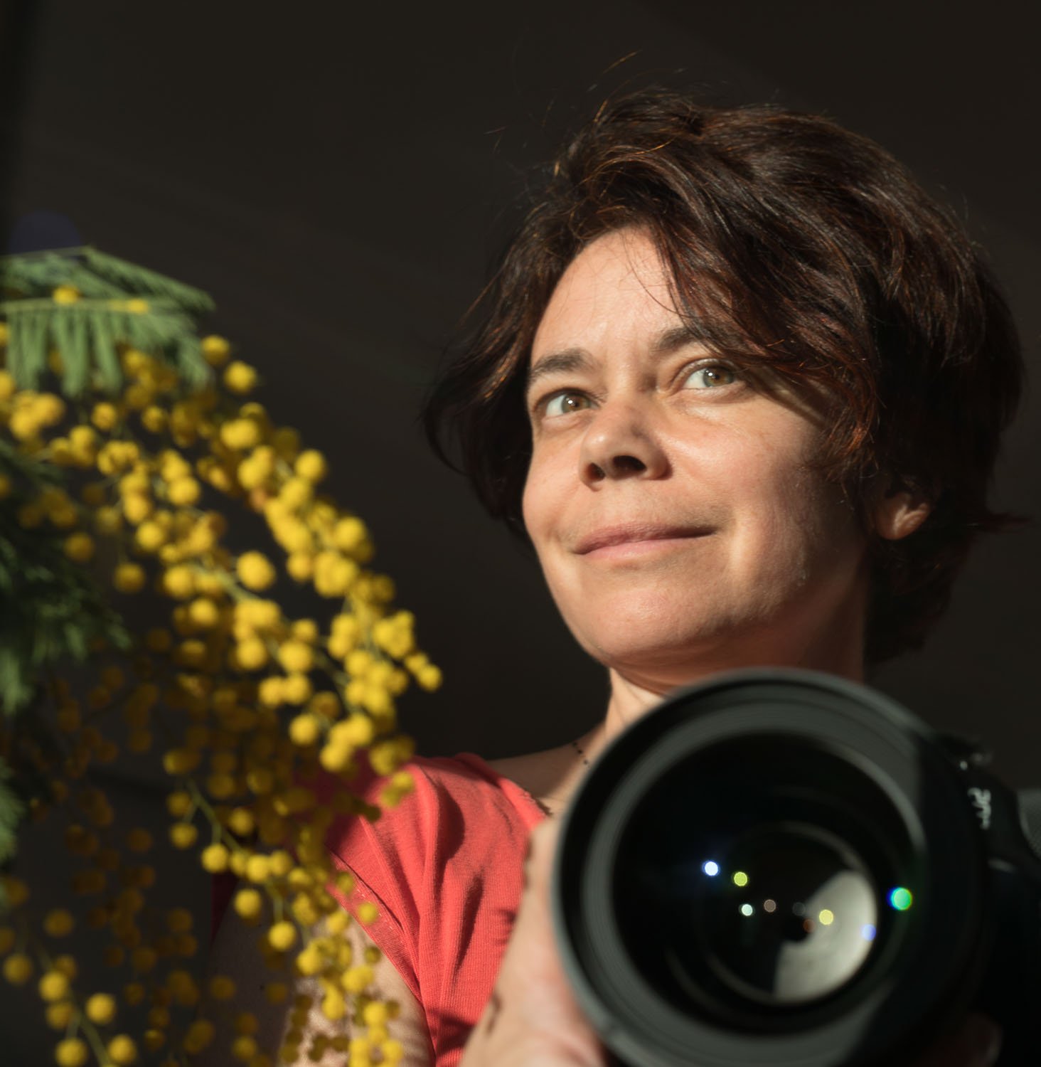 Auto-portrait de l'artiste photographe humaniste Magali Sacau. Visage éclairé et brin de Mimosa du Bassin d'Arcachon, l'appareil photo à la main.