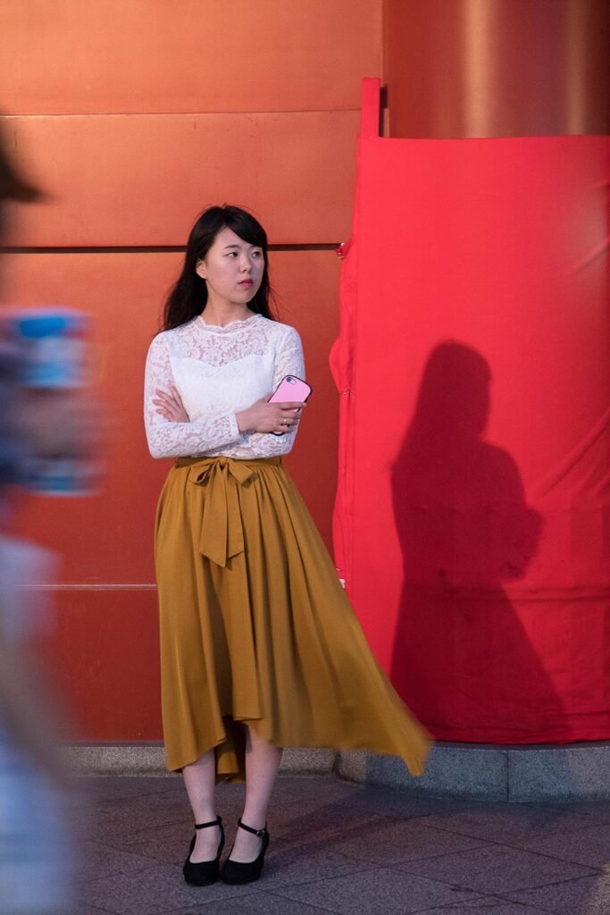 Cette photographie d’art éditée en tirages limités mise en vente, est le portrait d'une femme asiatique élégante et rêveuse. Cette œuvre poétique épurée sur fond de mur rouge qui contraste avec les touches de couleurs blanc, beige, et noir de la silhouette crée un effet très pictural. Cette photographie humaniste d'une élégante japonaise qui attend en rêvant dans le couloir du métro de Tokyo touchera les galeries comme les amateurs d'art par l'intemporalité de sa pause. Seule la présence de son téléphone portable à la main contraste avec l'intemporalité suggérée par la tenue en dentelle et la jupe fluide. Ce tableau photographique humaniste offrira l'évasion vers un ailleurs empli de douceur à son acquéreur, comme aux spectateurs lors d'une exposition.