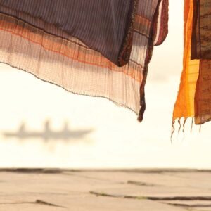 Étoffes roses orangées séchant au soleil sur les quais du Gange, en arrière plan une barque vogue avec trois silhouettes à bord. Une photographie d'art aux tonalités rose poudré et orangé. Lever de soleil sur le fleuve derrière des sarhis indiens étendus, très pictural et onirique. Douceur, sérénité, et rêves se dégagent de cette œuvre éditée sur papier fine art mise en vente en série limitée. Cette photographie artistique prise dans la cité sacrée de Bénarès, Varanasi séduira galeries d'art et collectionneurs. Une photo lumineuse et spirituelle qui emporte le spectateur vers un ailleurs divin empli de douceur.