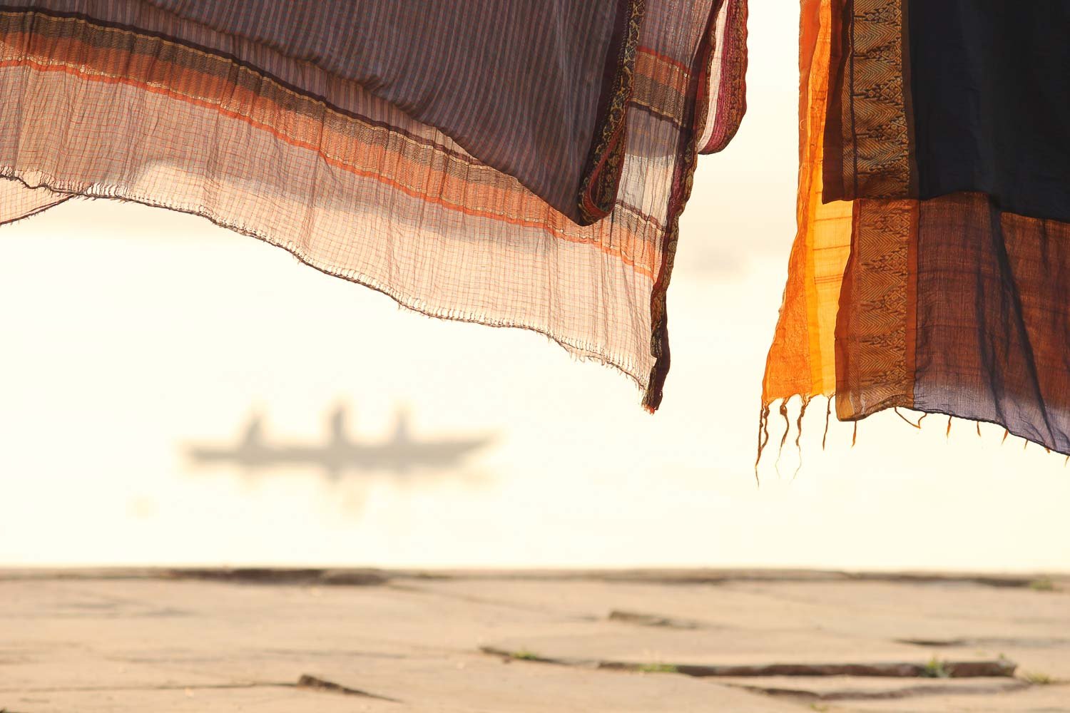 Étoffes roses orangées séchant au soleil sur les quais du Gange, en arrière plan une barque vogue avec trois silhouettes à bord. Une photographie d'art aux tonalités rose poudré et orangé. Lever de soleil sur le fleuve derrière des sarhis indiens étendus, très pictural et onirique. Douceur, sérénité, et rêves se dégagent de cette œuvre éditée sur papier fine art mise en vente en série limitée. Cette photographie artistique prise dans la cité sacrée de Bénarès, Varanasi séduira galeries d'art et collectionneurs. Une photo lumineuse et spirituelle qui emporte le spectateur vers un ailleurs divin empli de douceur.