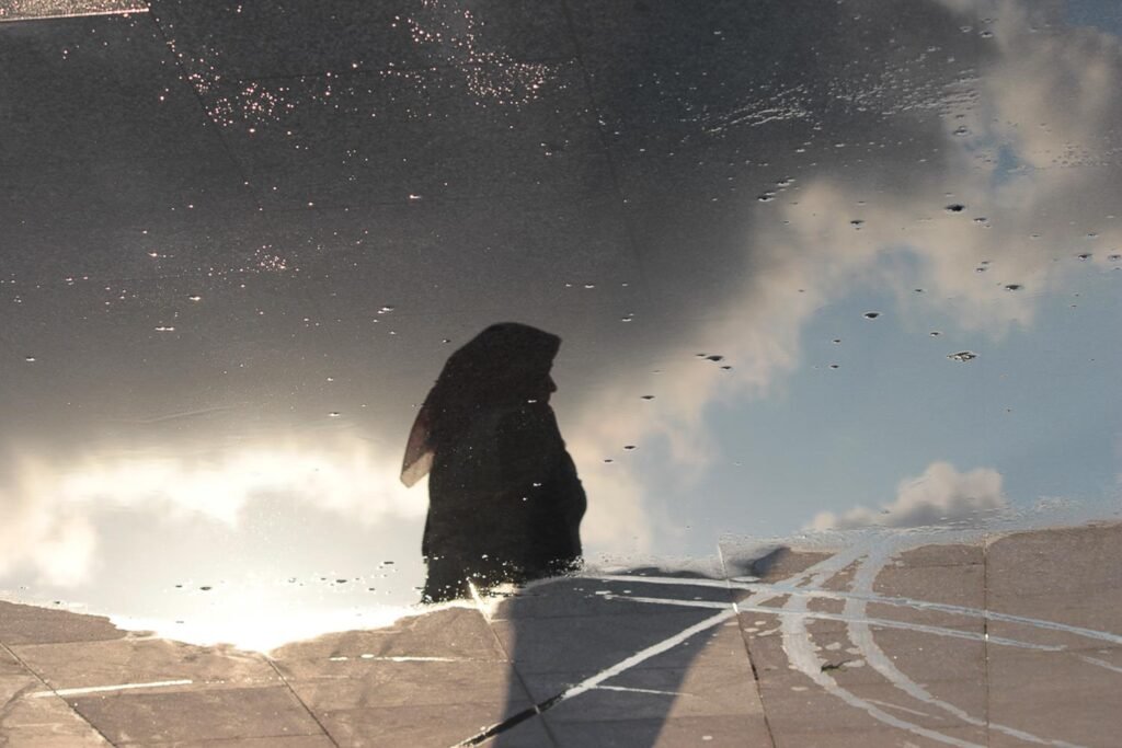 Photographie humaniste du Reflet d'une femme voilée devant un ciel nuageux dans une flaque d'eau sur le quai d'une station balnéaire en Turquie. Dans ce tirage d'art en édition limitée à dix exemplaires l'ombre noire se détache du ciel créant un contraste de couleurs onirique dans des nuances de gris. Les nuages orageux créent un ambiance inquiétante qui contraste avec la lumière profonde qui se dégage du ciel bleu. Cette œuvre d'art séduira les collectionneurs, comme les galeries en quête d'une photographie d'art contemporaine riche d'intemporalité, de rêve et d'évasion.